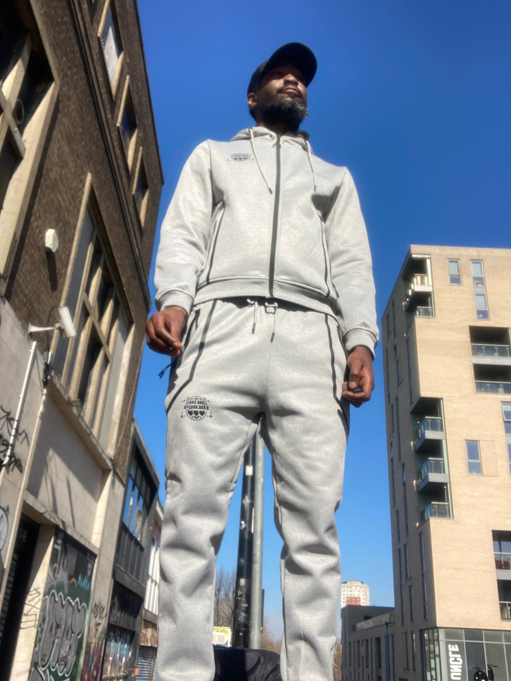 Person wearing a light gray tracksuit standing under a metal structure with a clear blue sky.